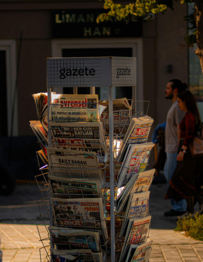 A newspaper stand with various publications on a sunny street corner.