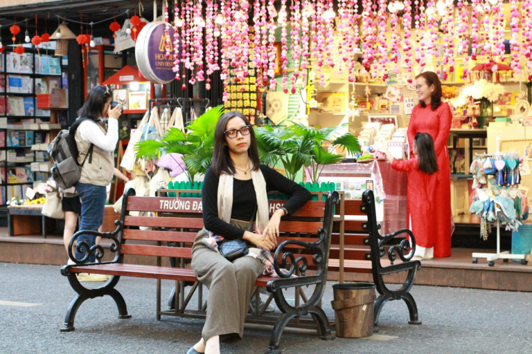 A woman sits on a bench in a lively Vietnamese market, surrounded by shoppers and colorful decorations.