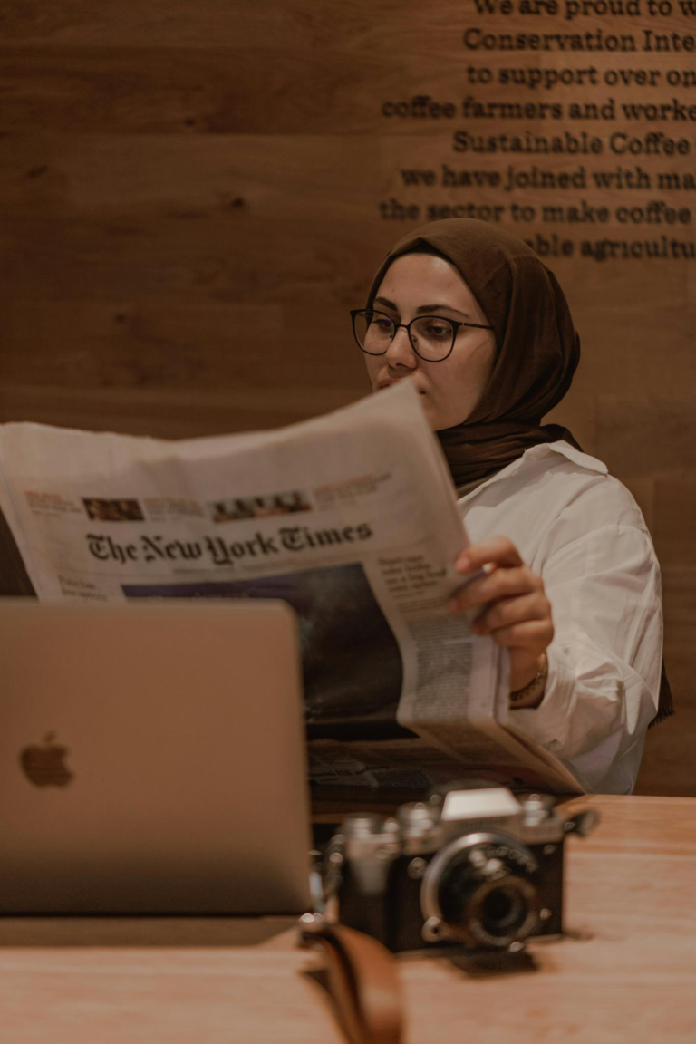 Muslim woman in hijab reading a newspaper at a café, laptop and camera on table.